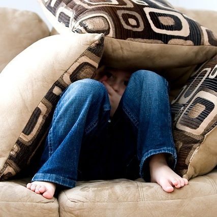 Kind versteckt sich zwischen Sofakissen – Sinnbild für ein geringes Selbstwertgefühl, das oft in der Kindheit entsteht